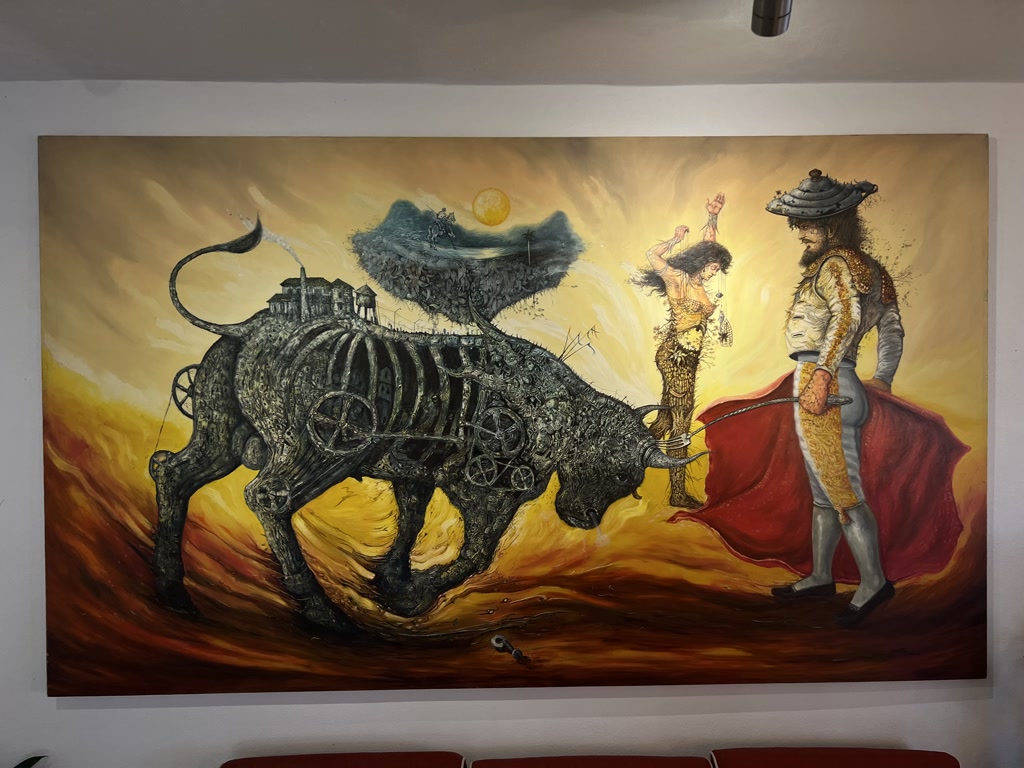 A large painting depicts a surreal scene with rich warm tones in the background, evoking a sunset. A mechanical bull dominates the left side of the piece, its body fashioned out of intricate machinery and wheels, with a transparent structure that reveals cogs and gears. Atop the bull, a small house and tree perch as if on a hill. A thin trail of smoke rises from the house's chimney, curling into the air. To the right, a matador stands in profile, donned in traditional bullfighting attire with embellishments, facing the mechanical bull. He holds a red cape behind him, suggesting a moment of tension with the bull. The matador's expression is focused, yet composed. Behind him stands a spectral figure resembling a flamenco dancer, seemingly made of wisps of light and shade. The dancer appears to be in motion, with one arm raised and the other holding what might be castanets. The overall tone of the painting is one of fantasy and drama, combining elements of Spanish culture with a steampunk aesthetic.