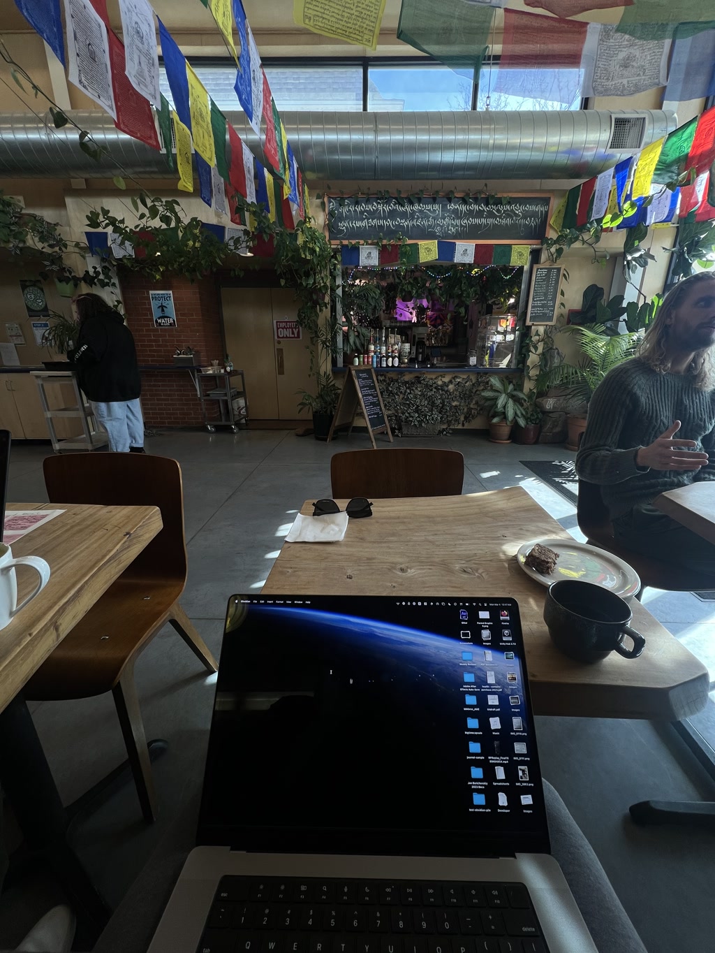 A cozy cafe setting with Tibetan prayer flags strung across the ceiling. Sunlight filters through large windows, casting a warm glow on the indoor plants and the rustic wooden furniture. A person stands at a counter in the back, while another person with long hair sits at a table in the foreground, partially blurred in motion. A laptop is open on a table in the lower right corner, its screen filled with numerous icons, next to a black coffee mug and a plate with a half-eaten treat. The colorful flags feature intricate designs and text, contributing to the serene and eclectic atmosphere.