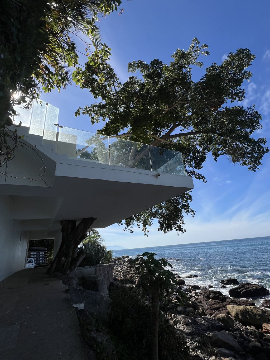 A modern overhanging structure with a glass balcony sits atop a support that blends into a rocky shoreline with the ocean in the background. The building is predominantly white and appears to be a contemporary design. A large, leafy tree emerges beside the building, its branches extending outward toward the sunlight. Beneath the structure, there is an open space leading towards the water, where waves can be seen hitting the rocks. A clear sky with minimal cloud cover suggests a bright, sunny day.