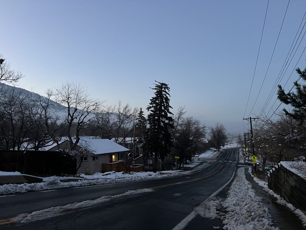 A wintery scene of a road after a snowfall with melting snow piled on the sides. Leafless trees line the road while evergreens add a touch of green. In the background, there's a hint of mountains partially shrouded by haze or low clouds. A house with snow on its roof and warm lighting visible through the window suggests a cozy atmosphere within. Street signs are present but the text is not legible. The sky is bright, hinting at early morning or late afternoon light.