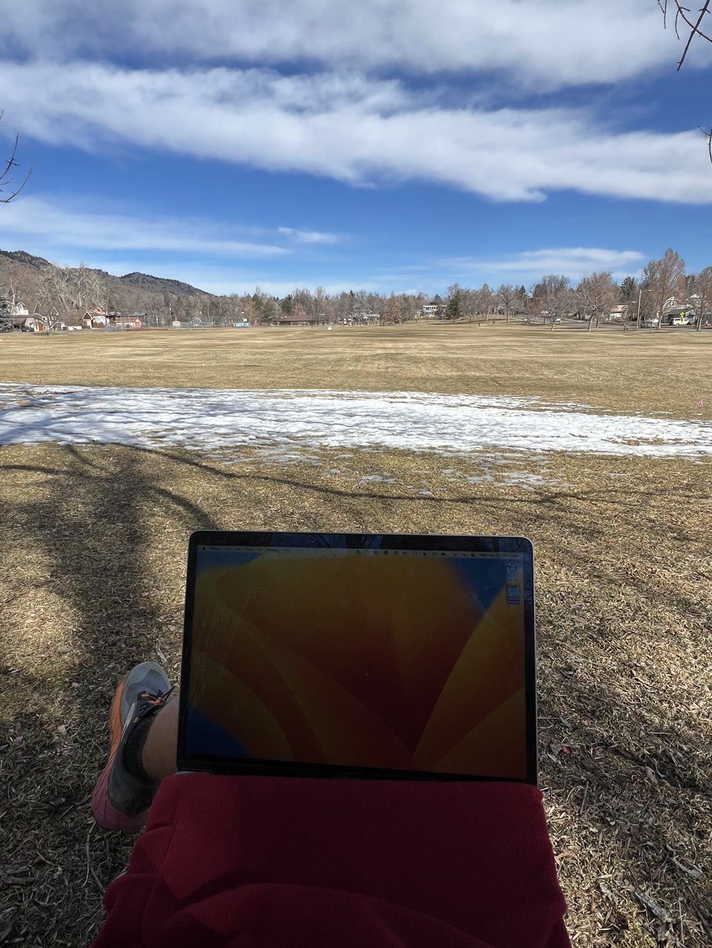 A person sits on a grassy field with remnants of snow scattered around, working on a laptop resting on their lap covered by a red piece of fabric, possibly a blanket. They are wearing a dark shoe and a portion of their leg is visible. The ground appears to be partly covered with shade, hinting at the presence of trees out of frame. Beyond the laptop is a vast open field edged by a line of trees and houses, with hills or low mountains visible in the background under a blue sky with some scattered white clouds.