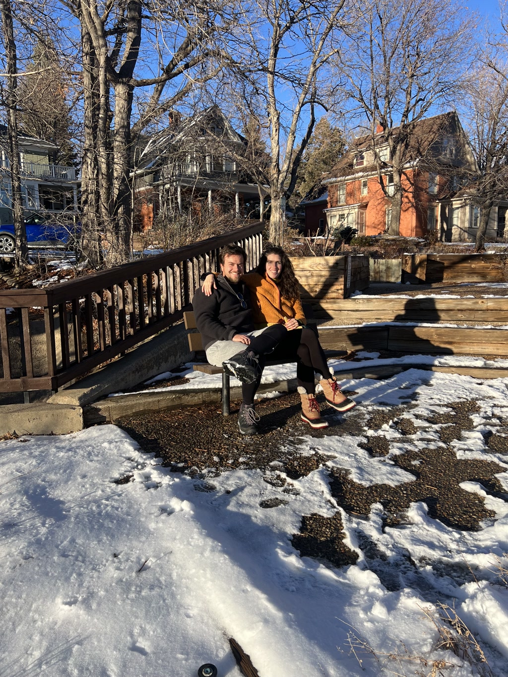 Two people are sitting on a wooden ledge flanked by bare trees, with residential buildings in the background. The ground and steps in the foreground are partially covered in snow, and there are clear patches where the snow has melted. The individuals appear to be enjoying a sunny day despite the cool surroundings suggested by their winter attire. The leaves on the trees are gone, indicating that it is either winter or late autumn. The male has casual clothing with a prominent watch on his left wrist and hands clasped, while the female is wearing a bright mustard-colored sweater, black leggings, and distinctive red boots with a fur trim. Both are smiling and seem to be in relaxed, cheerful spirits.