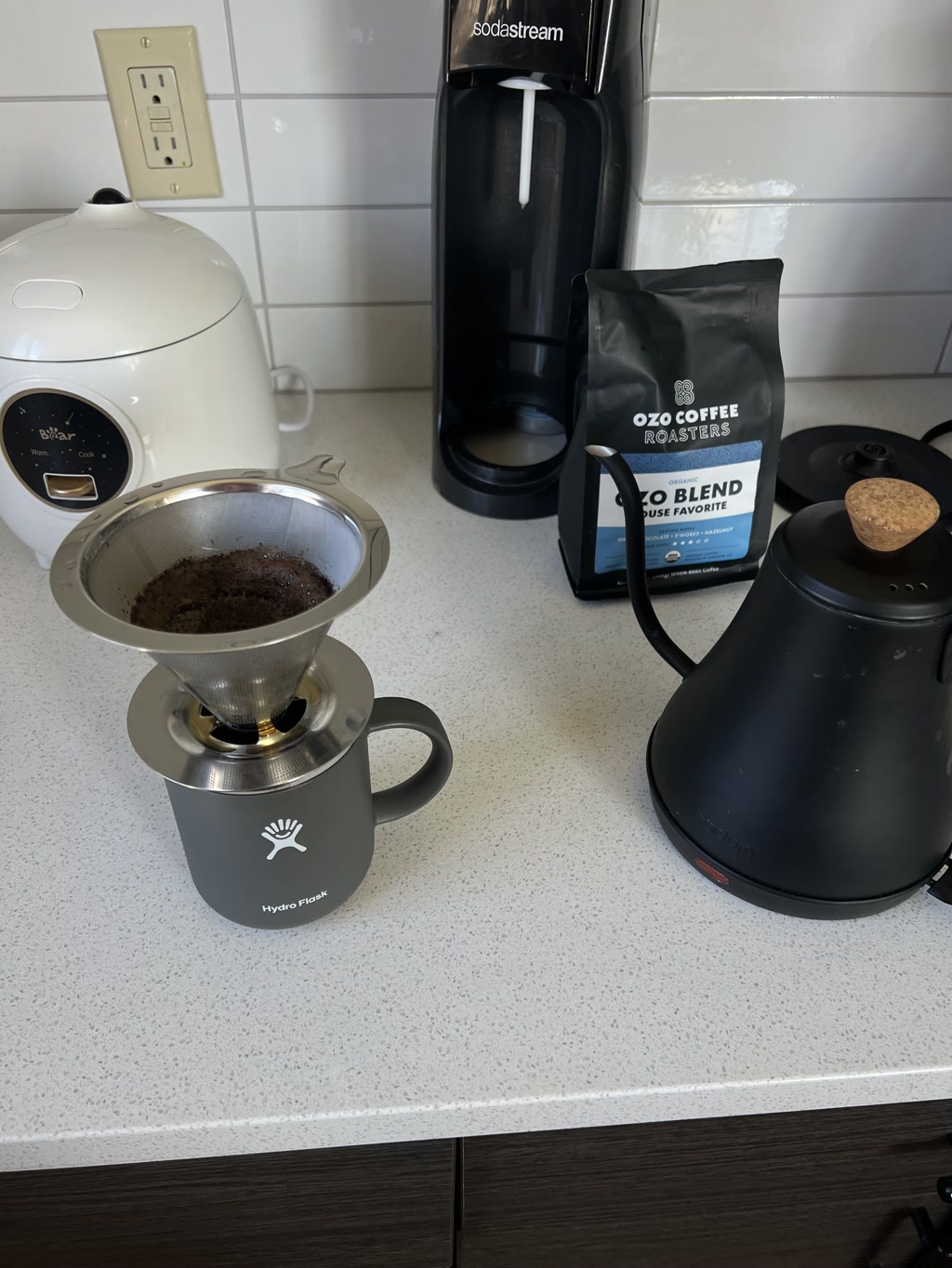 A kitchen countertop holding a variety of items used for making and serving coffee. On the left, there is a white rice cooker with a visible interface showing cooking options. In the middle, a SodaStream device stands next to a bag of coffee from OZO Coffee Roasters, advertising their 'OZO Blend' which is labeled as a 'House Favorite'. To the right, there is a kettle and in front is a Hydro Flask brand coffee mug with a metallic pour-over coffee dripper placed on top, filled with ground coffee.