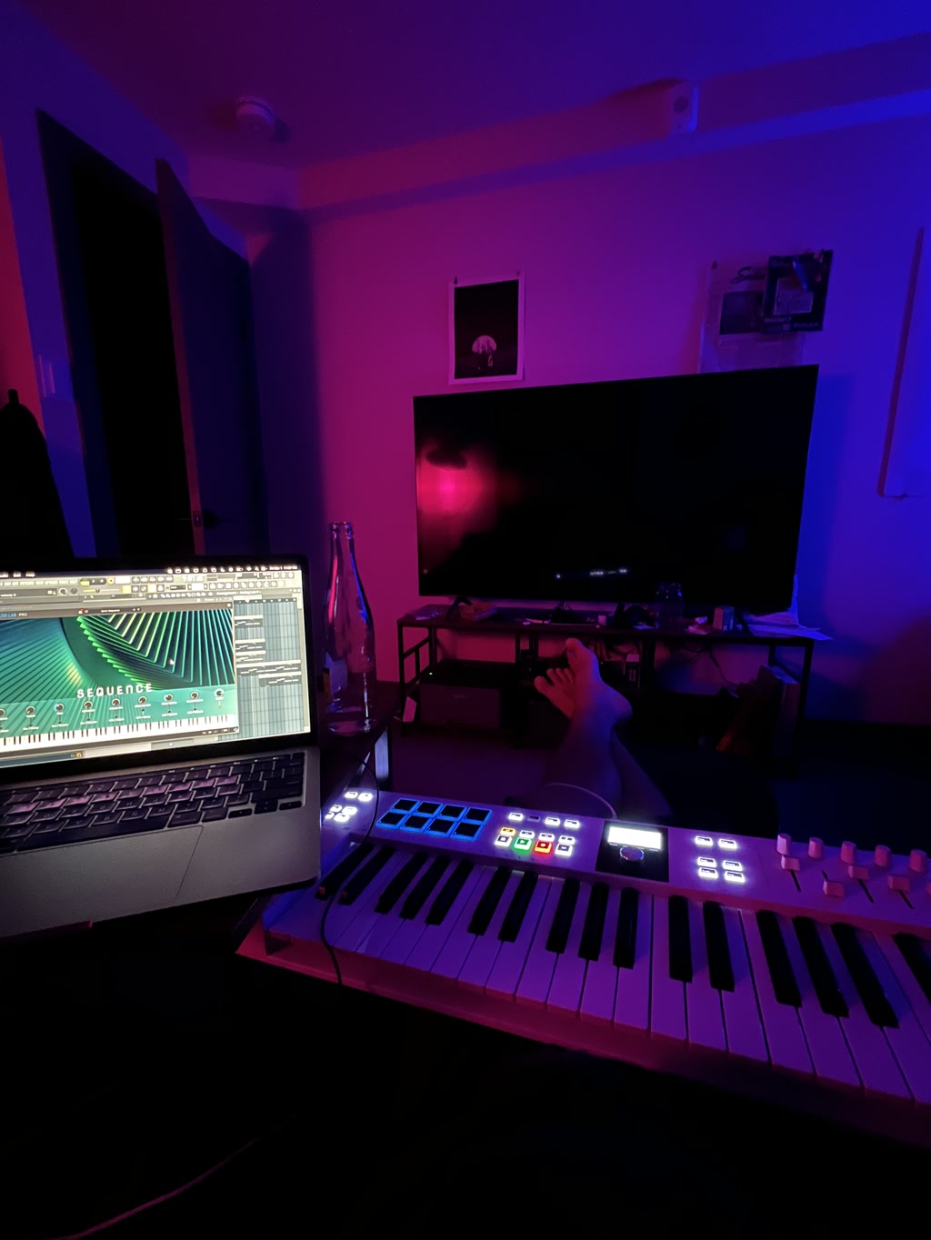 The setting is a dimly lit room with ambient lighting primarily in shades of blue and red. A laptop is open on a table with a music production software interface visible on its screen. In front of the laptop, there's a MIDI keyboard with illuminated pads and buttons. On the wall, there are two framed items, one appears to be a picture while the other is not clearly identifiable due to the lighting. There's a large flat-screen TV turned off on a stand across the room, and various items are scattered on the stand. The soft glow of multicolored lights gives the room a creative and relaxed atmosphere. There's a water bottle on the table and a person's feet are casually placed on an additional piece of furniture, indicating a comfortable, informal setting.