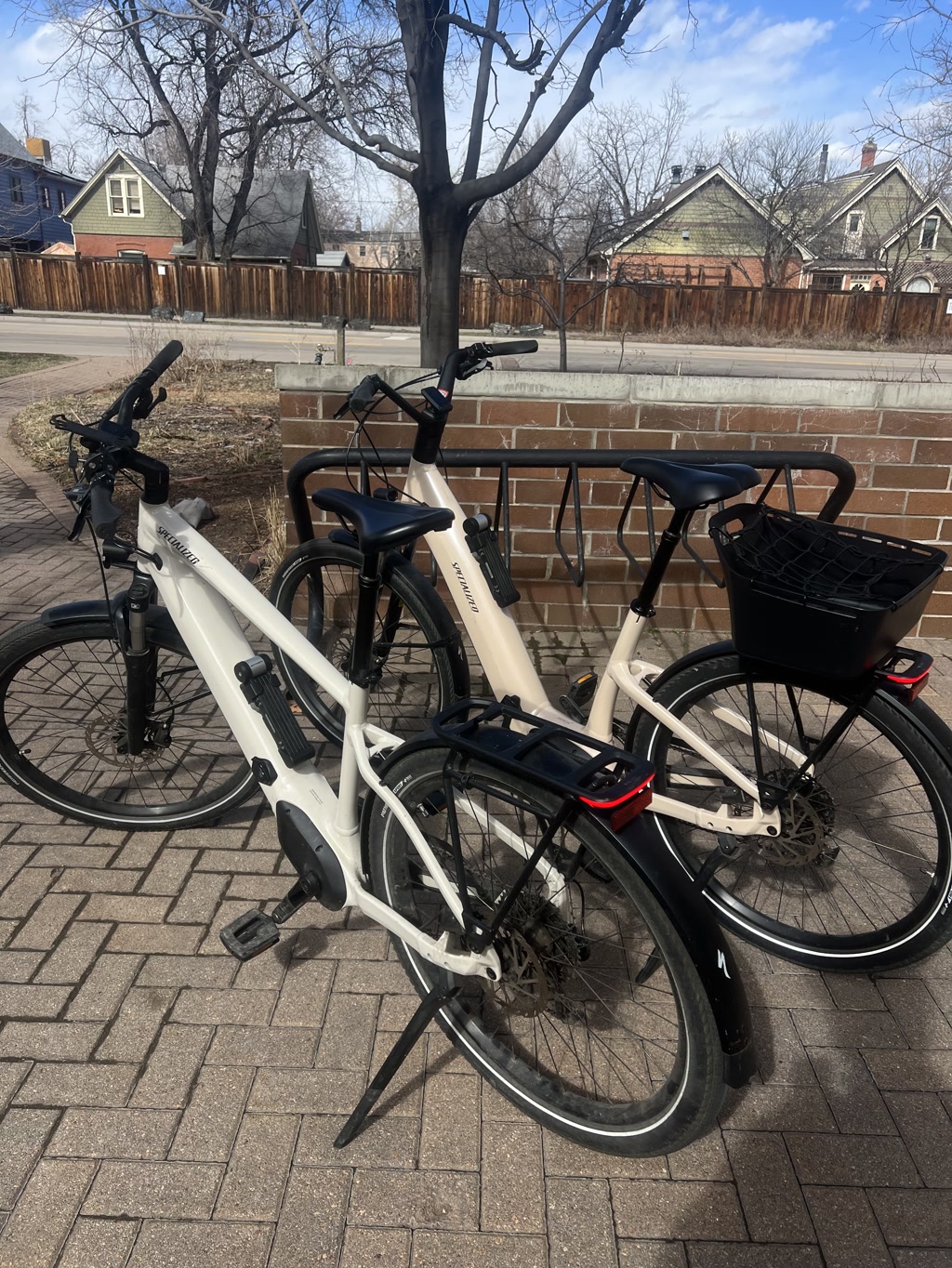 Two electric bicycles are parked side by side, locked to a bike rack. These bikes feature a sleek, modern design with primarily white frames accented by black components. Both are equipped with rear cargo racks, and one has a black basket attached to it. The handlebars show a clear design with ergonomic grips and straightforward controls, indicating a user-friendly interface. The setting is outdoors with a brick pavement, and in the background, there are bare trees and a wooden fence behind which residential houses can be seen.