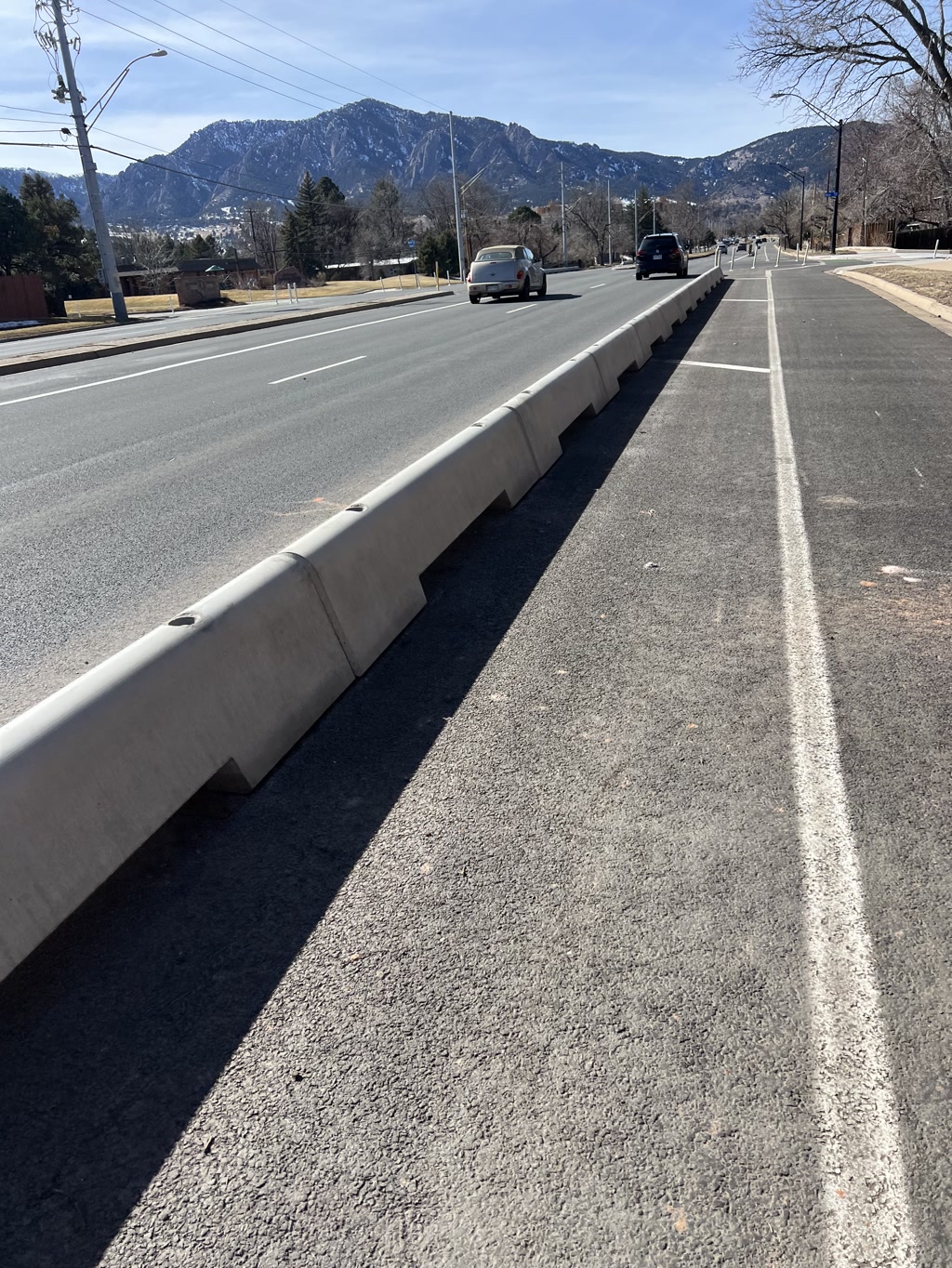 A roadway stretches into the distance with a prominent bike lane on the right-hand side, separated from the main road by a row of concrete barriers, ensuring cyclists' safety. Clear blue skies and a mountain range serve as the backdrop. The lanes are marked by white lines, and there are a few cars visible on the road. There is no text present.