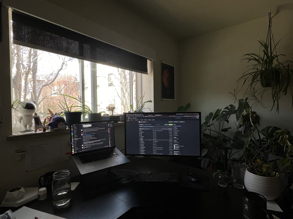 The scene shows a well-lit, cozy workspace featuring various plants beside and in front of a window with light filtering in. There's an evident focus on greenery, with multiple potted plants lending a calming, natural vibe to the environment. Two display monitors dominate the desk space, suggesting that the setup is used for work that requires extensive screen time, possibly coding or design. One of the monitors is mounted vertically, while the other is horizontal, flanking a laptop placed on a stand; all screens are on and display numerous lines of text and code, with a dark theme that is easier on the eyes. To the side, there's a glimpse of a poster or artwork on the wall. The desk also hosts miscellaneous items like a glasses case, a jar of water, a couple of coffee mugs, and what appears to be a small, white robot-like toy. Overall, the arrangement is practical yet personal, hinting at the occupant's preferences for technology, comfort, and a touch of nature.